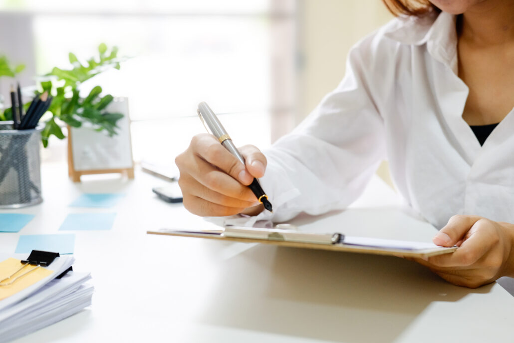 Close-up of a person in a white shirt writing on a clipboard at a bright desk with papers and plants.
