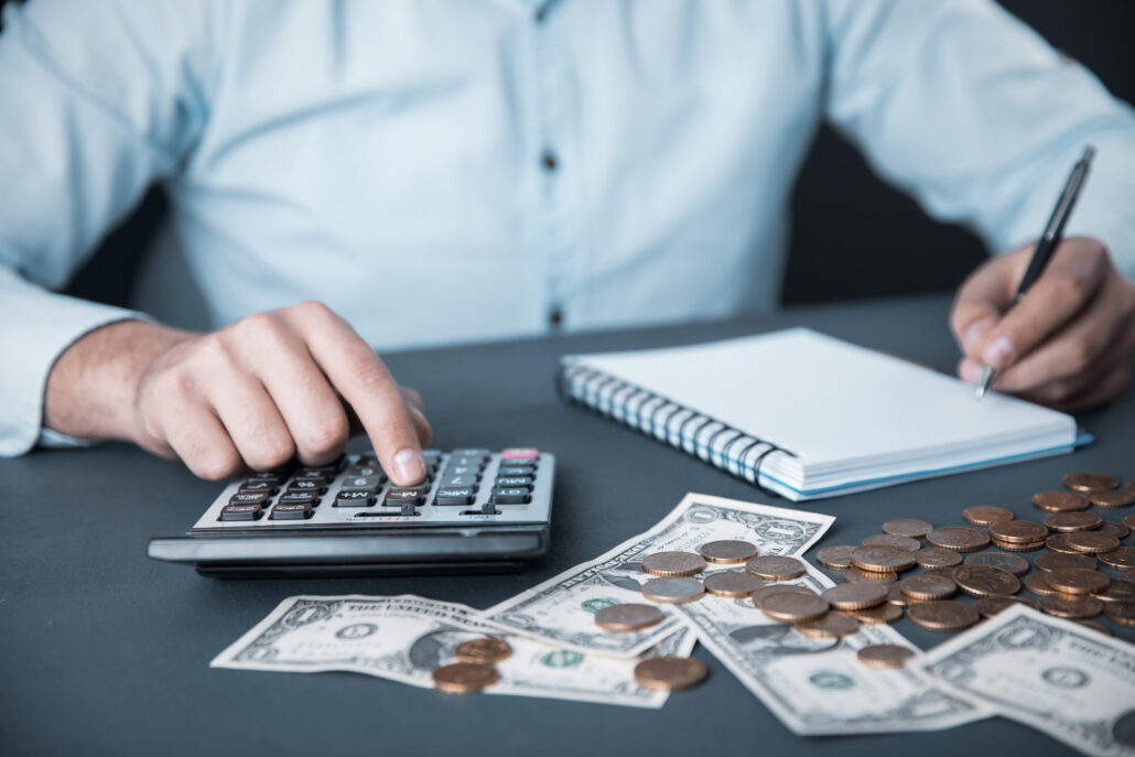 Business owner using a calculator with cash and coins on the desk while writing notes, representing determining reasonable compensation.