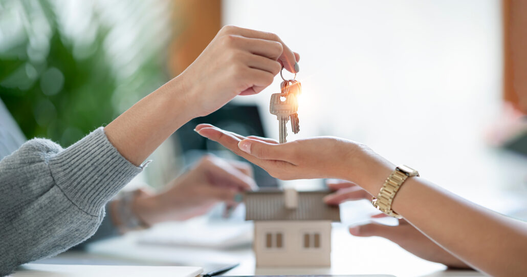 Person handing house keys to a buyer with a small model house on the table in the background