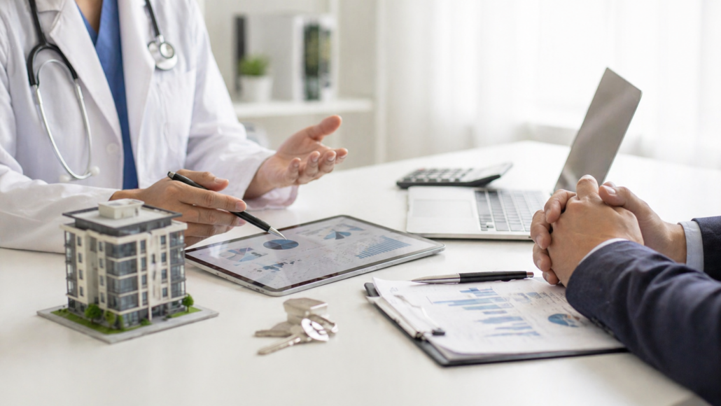 Doctor reviewing real estate tax planning charts with an advisor, property model, keys, calculator, and financial documents on a desk.